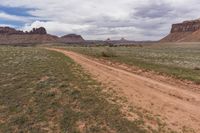 a dirt road with bushes on the side and tall rock formations in the background under a cloudy blue sky