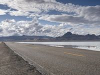 a lonely empty highway next to a body of water with mountains in the background, on a cloudy day