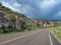 a highway going into the mountains with dark clouds overhead it is surrounded by some rocks