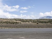 a motorcycle riding on the street beside a hill on a sunny day with cloudy sky