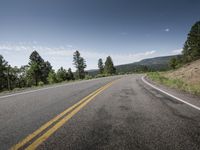 an empty road next to the mountains and trees with some clouds in the sky above