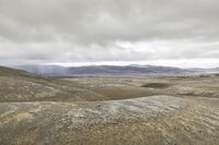 a lone person is walking through the barren hills on a cloudy day, and there are mountains in the distance