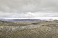 a lone person is walking through the barren hills on a cloudy day, and there are mountains in the distance