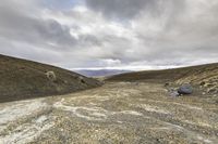 a barren area with rocks and dirt on the ground with mountains in the distance and clouds