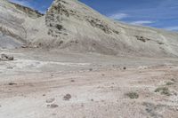 a dirt field with a dirt mountain in the distance and some rocks, bushes, and sparse grass