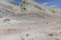 a dirt field with a dirt mountain in the distance and some rocks, bushes, and sparse grass