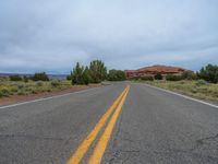 a empty long road with a yellow line painted down the middle of the street and in between two bushes, a red rock formation is visible in the background