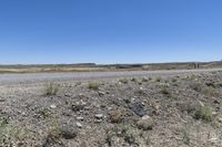 a rural landscape, with small rocks and scrubland, and an open area with large buildings and a few cars parked on the side