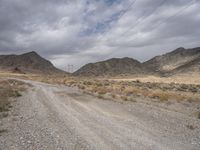 gravel road leading through barren mountains with telephone poles and power lines in the background and a grey cloudy sky