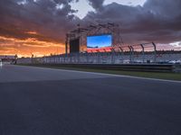 a television screen in the sky by a track with cars driving around it at sunset