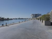 an empty walkway beside a waterway with several parked boats on it and a glass building behind