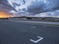 a street lined with white markers under a cloudy sky at sunset on a runway with cars in the background