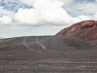 a large open dirt area with some red hills and clouds in the distance, and a person on a horse standing at the end of the path