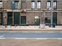 a brick building with green doors, sidewalk and bike leaning against it and some garbage containers in front of the buildings