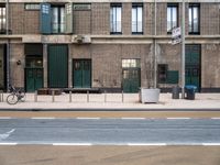 a brick building with green doors, sidewalk and bike leaning against it and some garbage containers in front of the buildings