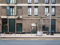 a brick building with green doors, sidewalk and bike leaning against it and some garbage containers in front of the buildings