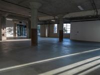 an empty warehouse has a wall of windows behind it in this area, a concrete floor with cement columns, and flooring are exposed