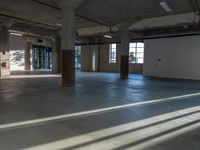 an empty warehouse has a wall of windows behind it in this area, a concrete floor with cement columns, and flooring are exposed