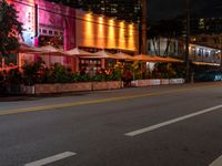 a city road and sidewalk filled with tables and chairs at night with bright lights on the buildings