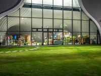 a glass building with a lawn and walkways between two buildings at night that have the lights on and the reflection in the window