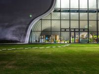 a glass building with a lawn and walkways between two buildings at night that have the lights on and the reflection in the window