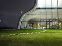 a glass building with a lawn and walkways between two buildings at night that have the lights on and the reflection in the window