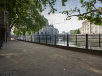 a bench that is by a fence in the sidewalk looking at a river and buildings