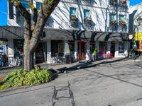 a couple of buildings on a city street corner with flowers hanging on the windows, plants on boxes and trees along side of road