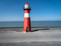 the lighthouse has a red and white color on its base near the ocean, and is next to a parking lot