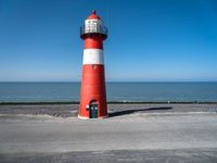 the lighthouse has a red and white color on its base near the ocean, and is next to a parking lot