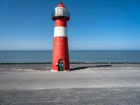 the lighthouse has a red and white color on its base near the ocean, and is next to a parking lot