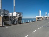 view of industrial structures, with sea and blue sky in background, from parking area, on sidewalk near roadway