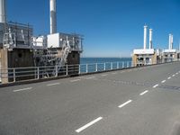 view of industrial structures, with sea and blue sky in background, from parking area, on sidewalk near roadway