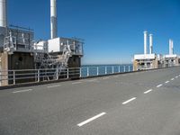 view of industrial structures, with sea and blue sky in background, from parking area, on sidewalk near roadway