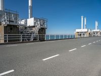 view of industrial structures, with sea and blue sky in background, from parking area, on sidewalk near roadway