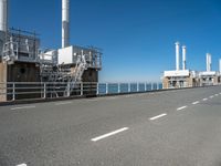 view of industrial structures, with sea and blue sky in background, from parking area, on sidewalk near roadway