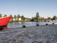 a large red boat sitting on top of a brick road near water and trees at its side