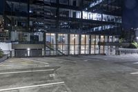 an empty parking lot in front of a building at night with buildings behind it and stairs