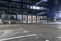 an empty parking lot in front of a building at night with buildings behind it and stairs