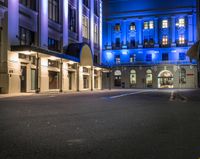 an empty street lined with tall buildings under night lights, and there are blue lights on the building windows