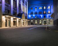 an empty street lined with tall buildings under night lights, and there are blue lights on the building windows