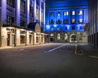 an empty street lined with tall buildings under night lights, and there are blue lights on the building windows