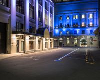 an empty street lined with tall buildings under night lights, and there are blue lights on the building windows