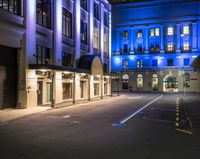 an empty street lined with tall buildings under night lights, and there are blue lights on the building windows