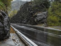 a wet asphalt road in the forest, with rock walls to the side and a man riding a bicycle in the background