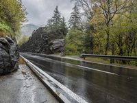 a wet asphalt road in the forest, with rock walls to the side and a man riding a bicycle in the background