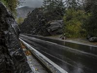 a wet asphalt road in the forest, with rock walls to the side and a man riding a bicycle in the background