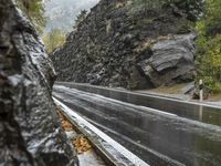 a wet asphalt road in the forest, with rock walls to the side and a man riding a bicycle in the background
