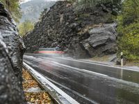 a wet asphalt road in the forest, with rock walls to the side and a man riding a bicycle in the background