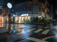 a wet city street in the rain at night with a store with shops and plants in the windows